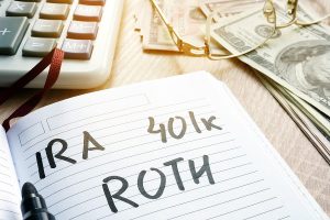 A notebook with "IRA," "401k," and "ROTH" written on it, surrounded by cash, a calculator, and glasses.
