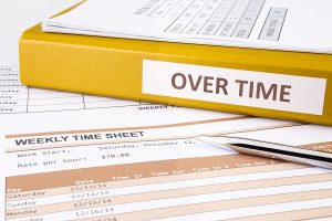 A yellow binder labeled "Overtime" on top of weekly timesheets and a pen, representing overtime pay exemptions and payroll management.